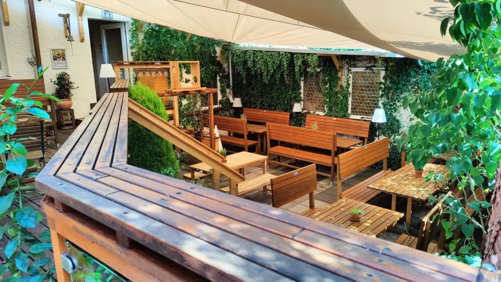 Biergarten des Atelier Teufelsbäck mit Sonnensegeln, umfassender Bepflanzung und hölzernen Stühlen und Bänken.