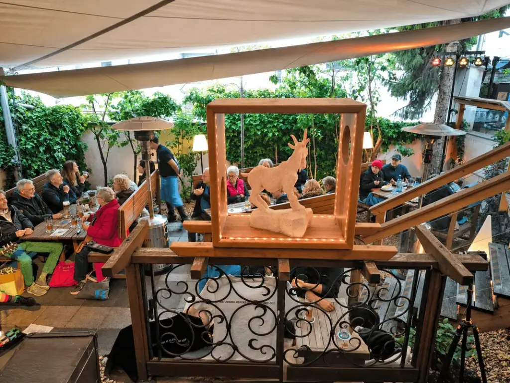 Blick auf den gefüllten Biergarten - im Vordergrund das metallene Tor, darüber die Ton-Ziege, welches das Maskottchen des Atelier Teufelsbäck ist.